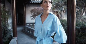 Woman wearing a blue striped belted shirt dress, standing in a serene courtyard, exuding effortless sophistication and timeless style.