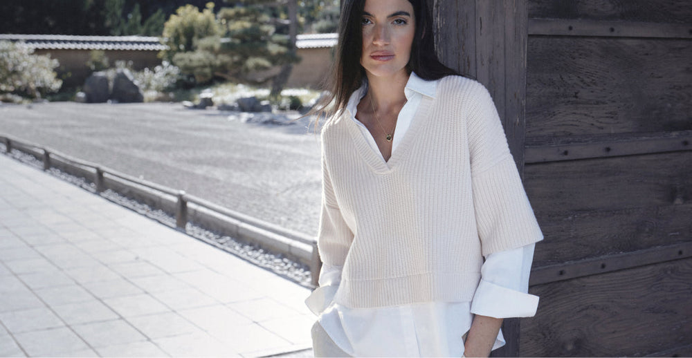 Woman wearing a cream knit V-neck sweater layered over a white button-down shirt, styled effortlessly in an outdoor setting with natural light.