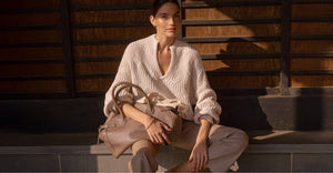 Woman in a knit sweater holding a taupe handbag, seated on steps with warm wooden panels in the background.